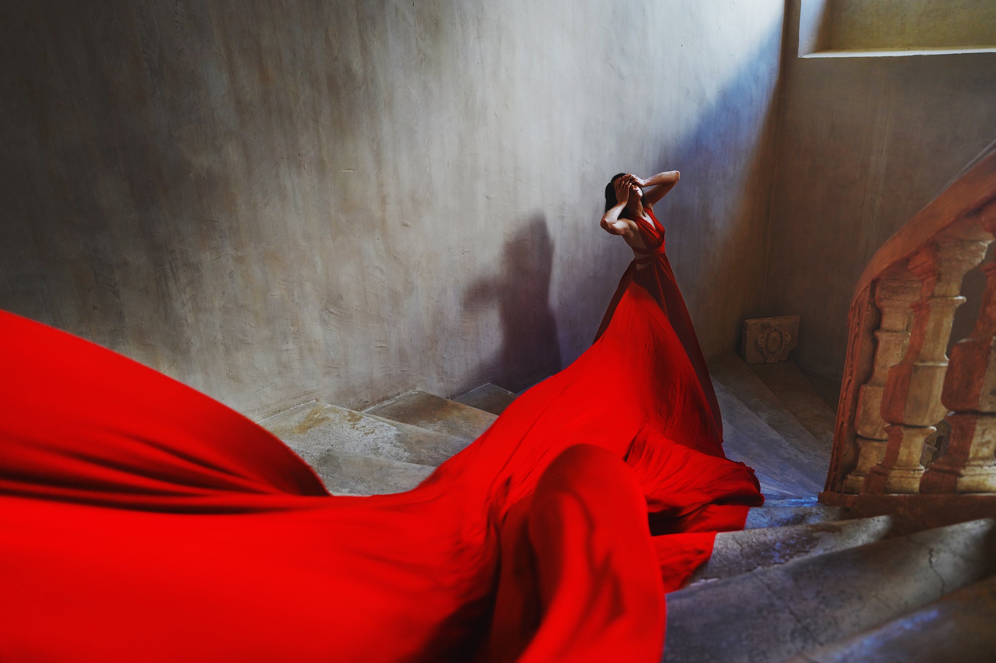 A woman in a dramatic red gown stands at the top of a stone staircase, hands covering her face, as her voluminous red skirt cascades down the steps in a sweeping flow against a textured grey wall.
