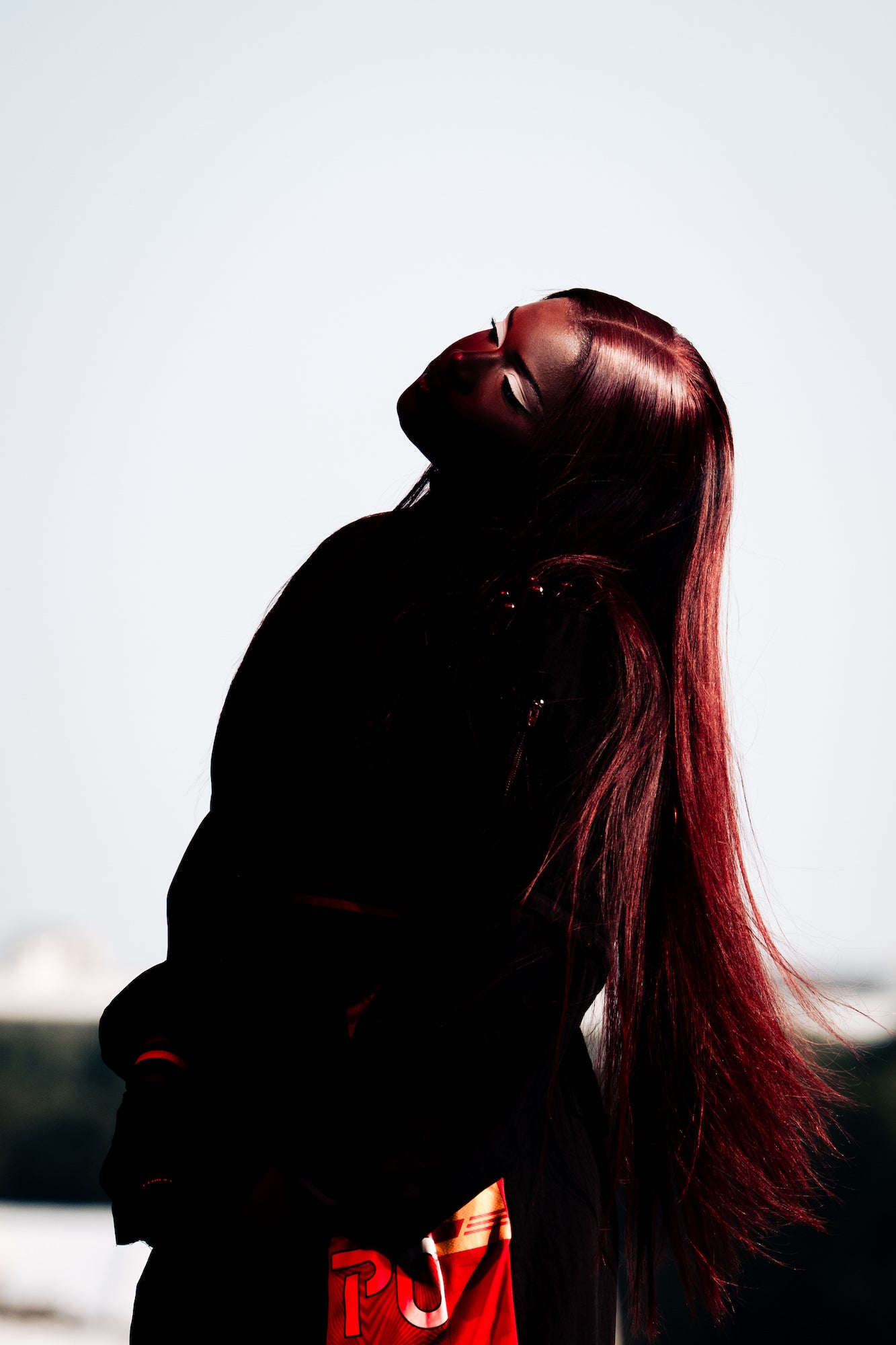 Backlit portrait with long auburn hair; head tilted, dramatic contrast against sky. Backlit portrait with long auburn hair; head tilted, dramatic contrast against sky.
