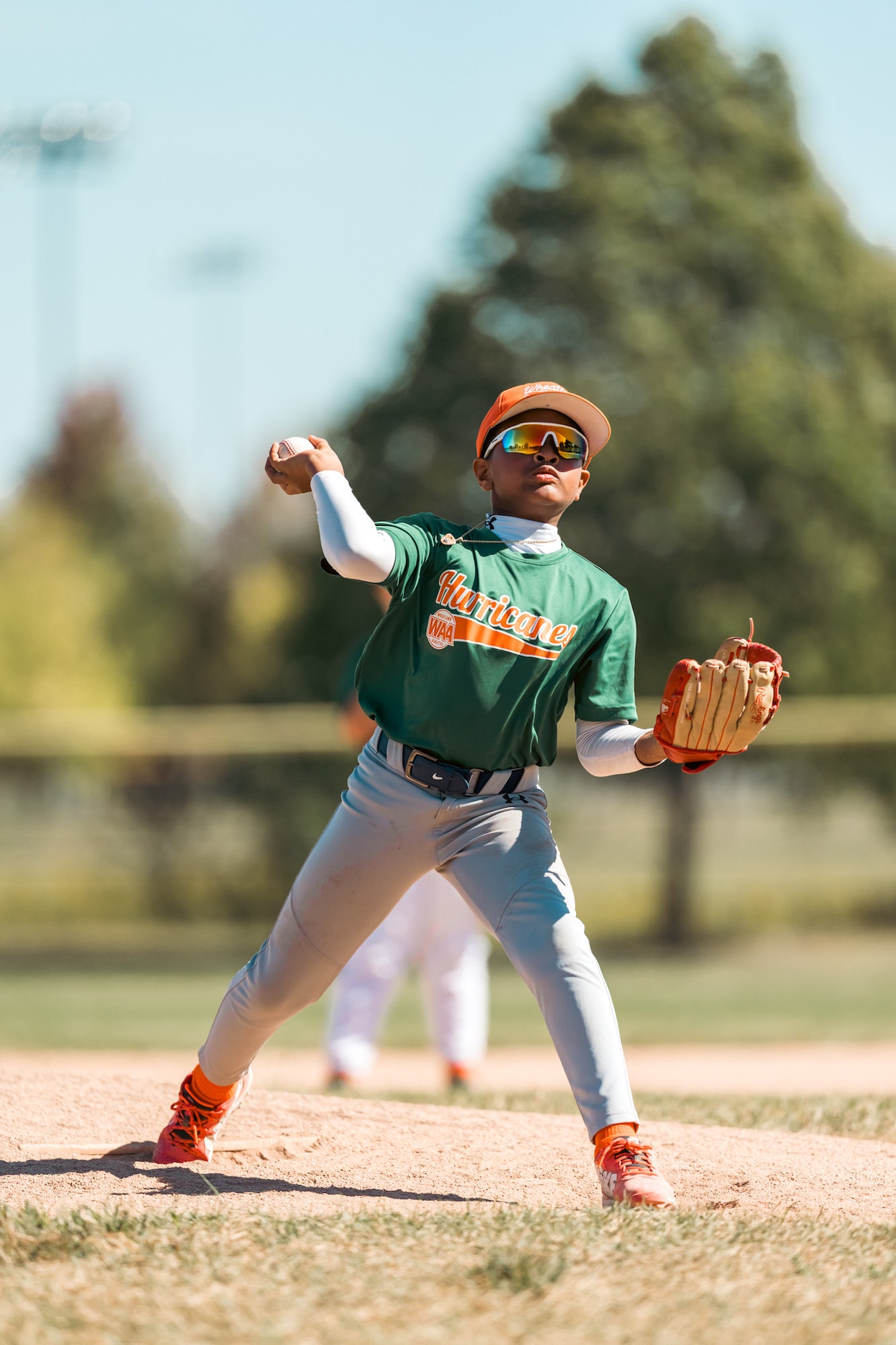 Youth pitcher in green uniform fires from the mound, sunglasses gleaming in bright spring light. Youth pitcher in green uniform fires from the mound, sunglasses gleaming in bright spring light.