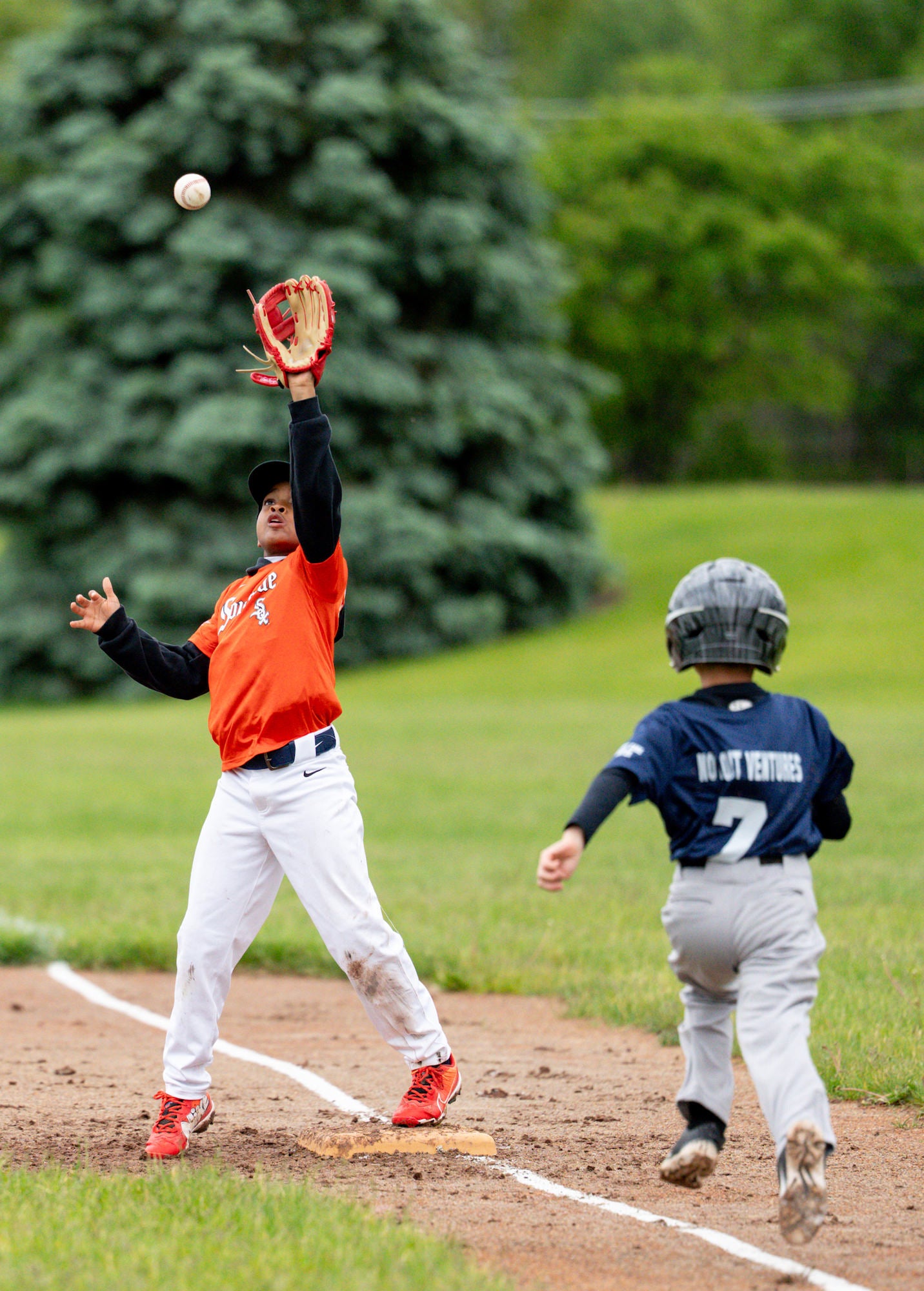 First baseman reaches high for a throw at the bag as the runner charges down the line. First baseman reaches high for a throw at the bag as the runner charges down the line.