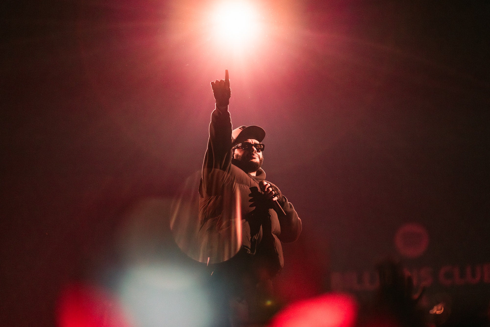 Performer points toward a bright stage light, bathed in warm red glow. Performer points toward a bright stage light, bathed in warm red glow.