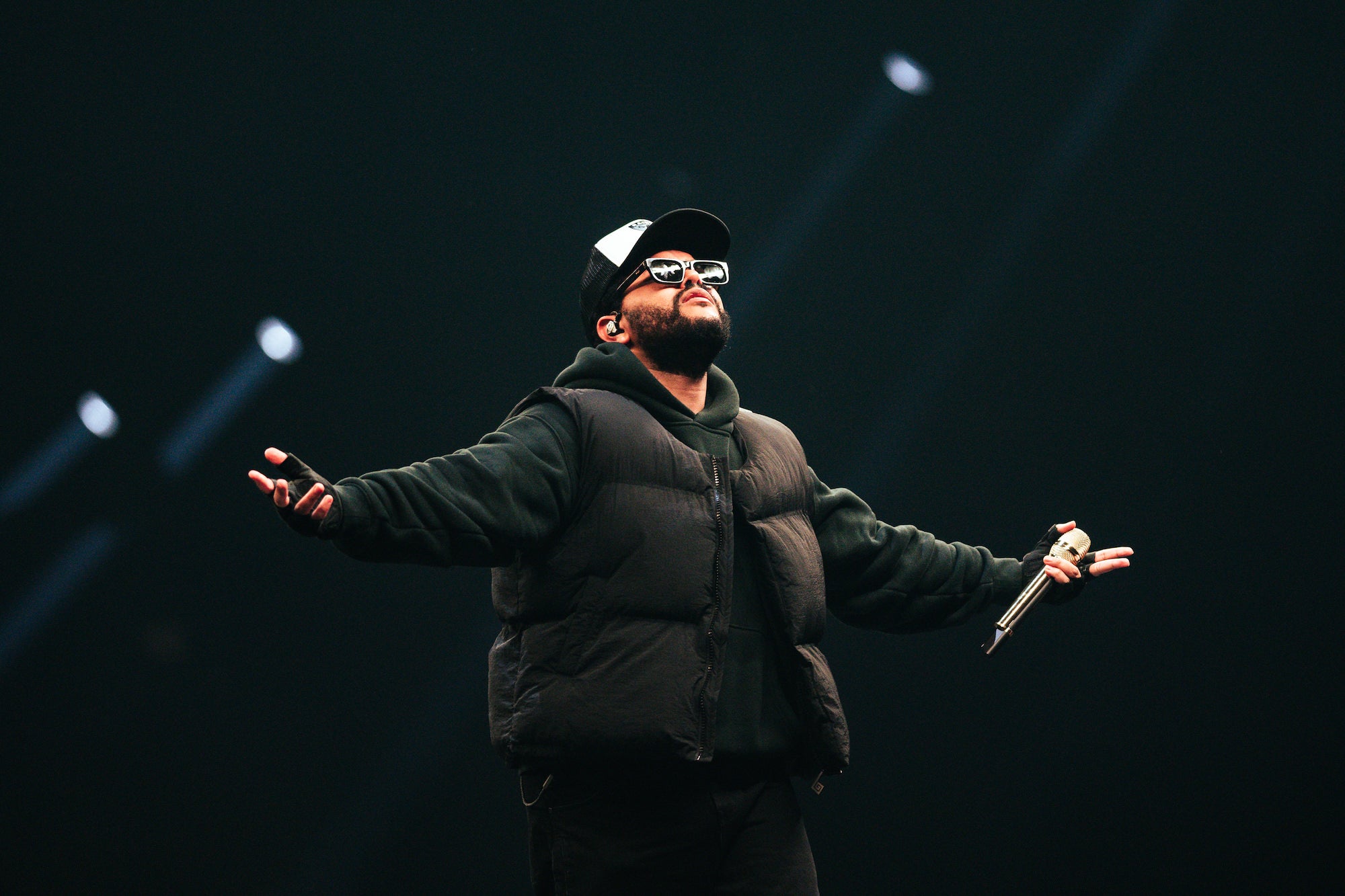 Performer in dark vest and cap stands arms wide on a dim stage, holding a mic, spotlights cutting through the dark. Performer in dark vest and cap stands arms wide on a dim stage, holding a mic, spotlights cutting through the dark.