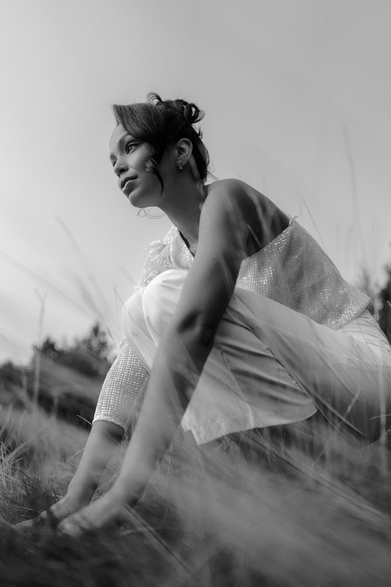 Low-angle black-and-white portrait; person crouching in tall grass, thoughtful gaze. Low-angle black-and-white portrait; person crouching in tall grass, thoughtful gaze.