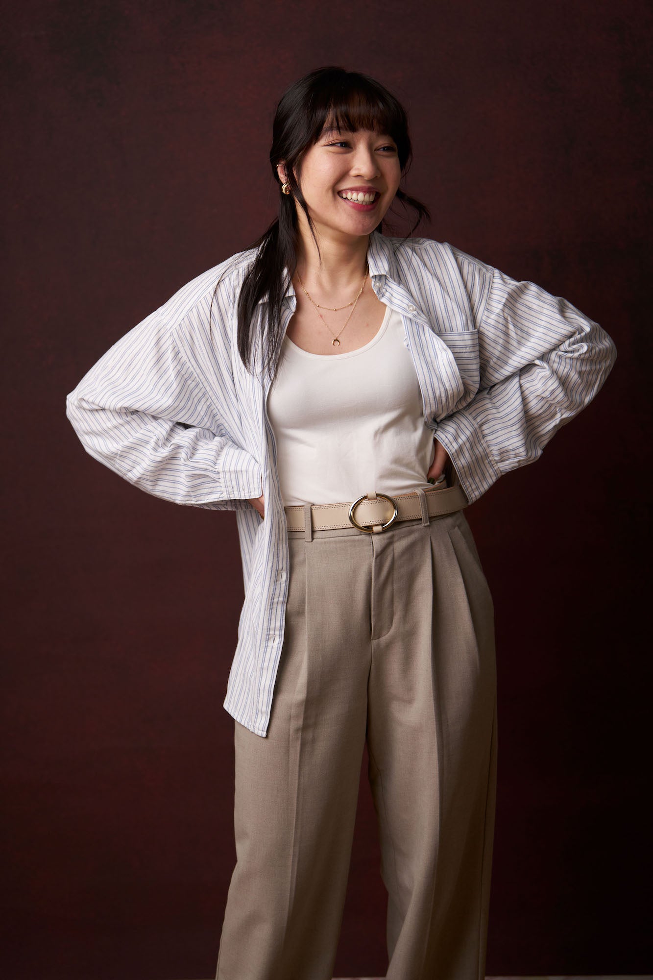 Full-length portrait of a woman laughing with hands on hips, striped oversized shirt and beige trousers, warm studio background. Full-length portrait of a woman laughing with hands on hips, striped oversized shirt and beige trousers, warm studio background.