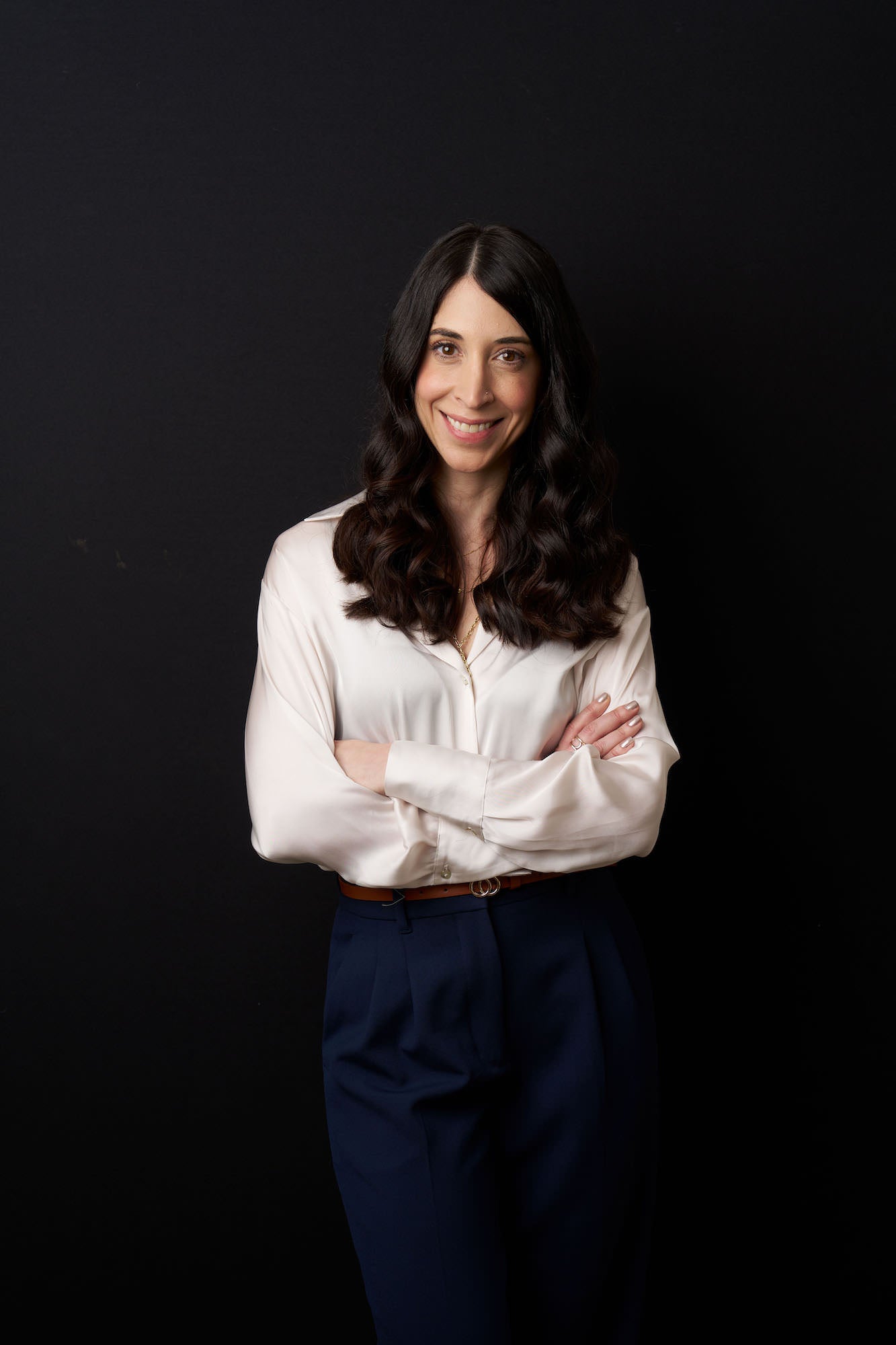 Three-quarter studio portrait of a woman in a cream blouse and navy trousers, arms crossed against a black backdrop. Three-quarter studio portrait of a woman in a cream blouse and navy trousers, arms crossed against a black backdrop.