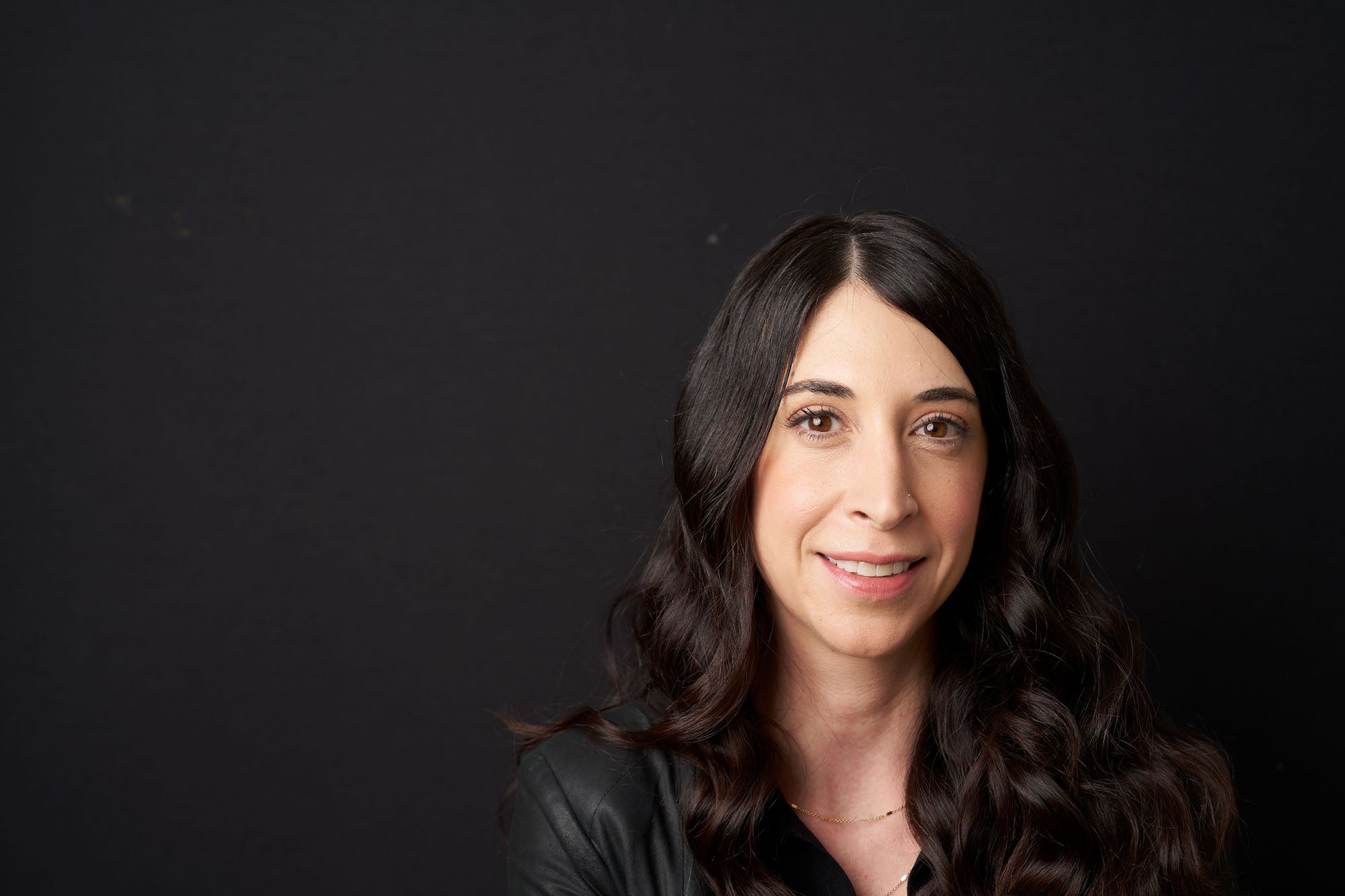 Studio headshot of a woman with long dark wavy hair against a black backdrop, softly lit and smiling. Studio headshot of a woman with long dark wavy hair against a black backdrop, softly lit and smiling.