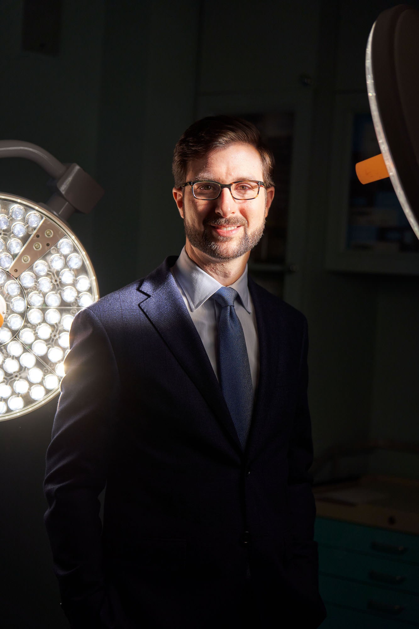 Professional portrait of a man in a suit and glasses lit by circular surgical lights in a clinical setting. Professional portrait of a man in a suit and glasses lit by circular surgical lights in a clinical setting.
