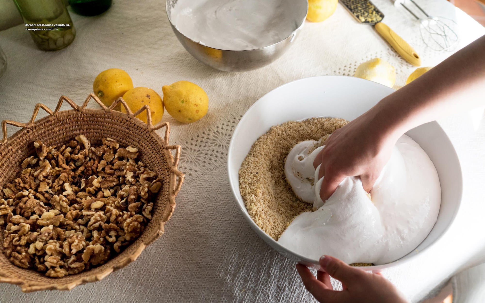 Hands fold fluffy meringue into ground nuts beside lemons and a basket of walnuts on a kitchen table.