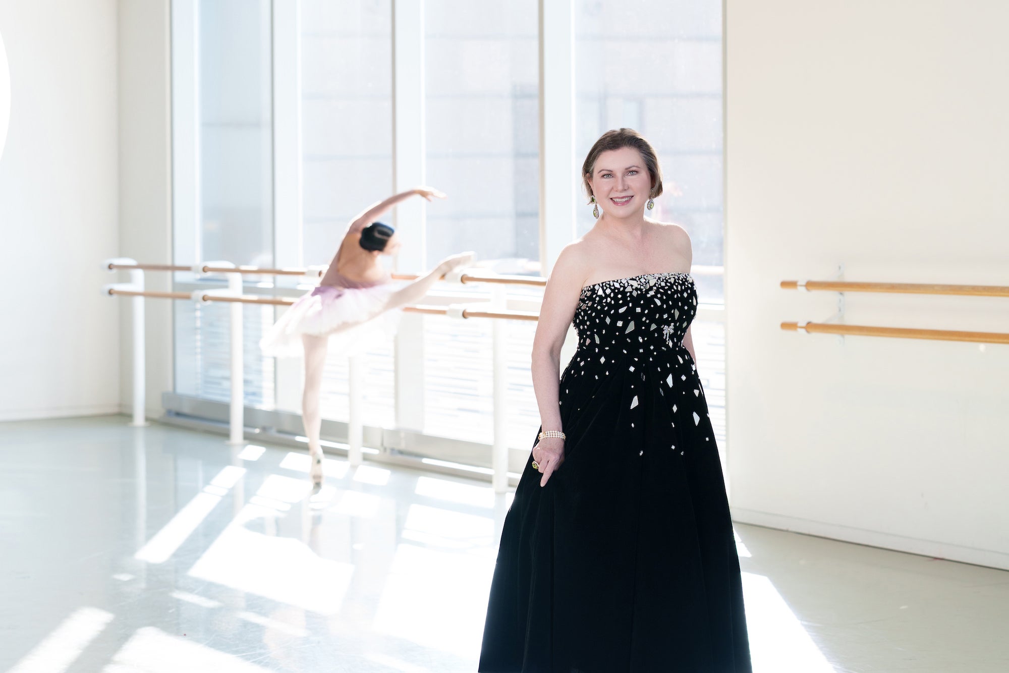 Woman in a black strapless gown in a bright ballet studio; dancer blurred in the background at the barre.